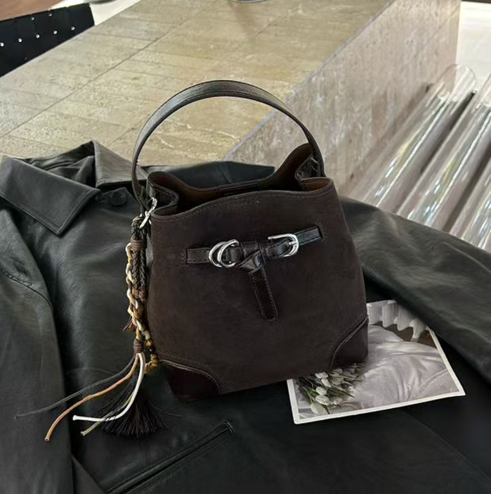 Brown Briar Bucket Bag with a curved top handle, silver-tone buckle details and a tassel charm, resting on a dark jacket.